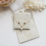 A necklace and earrings set are displayed on a white surface, accompanied by a small white flower and a beige pouch with the text "Sarah BRIDGETSmaid" printed on it.