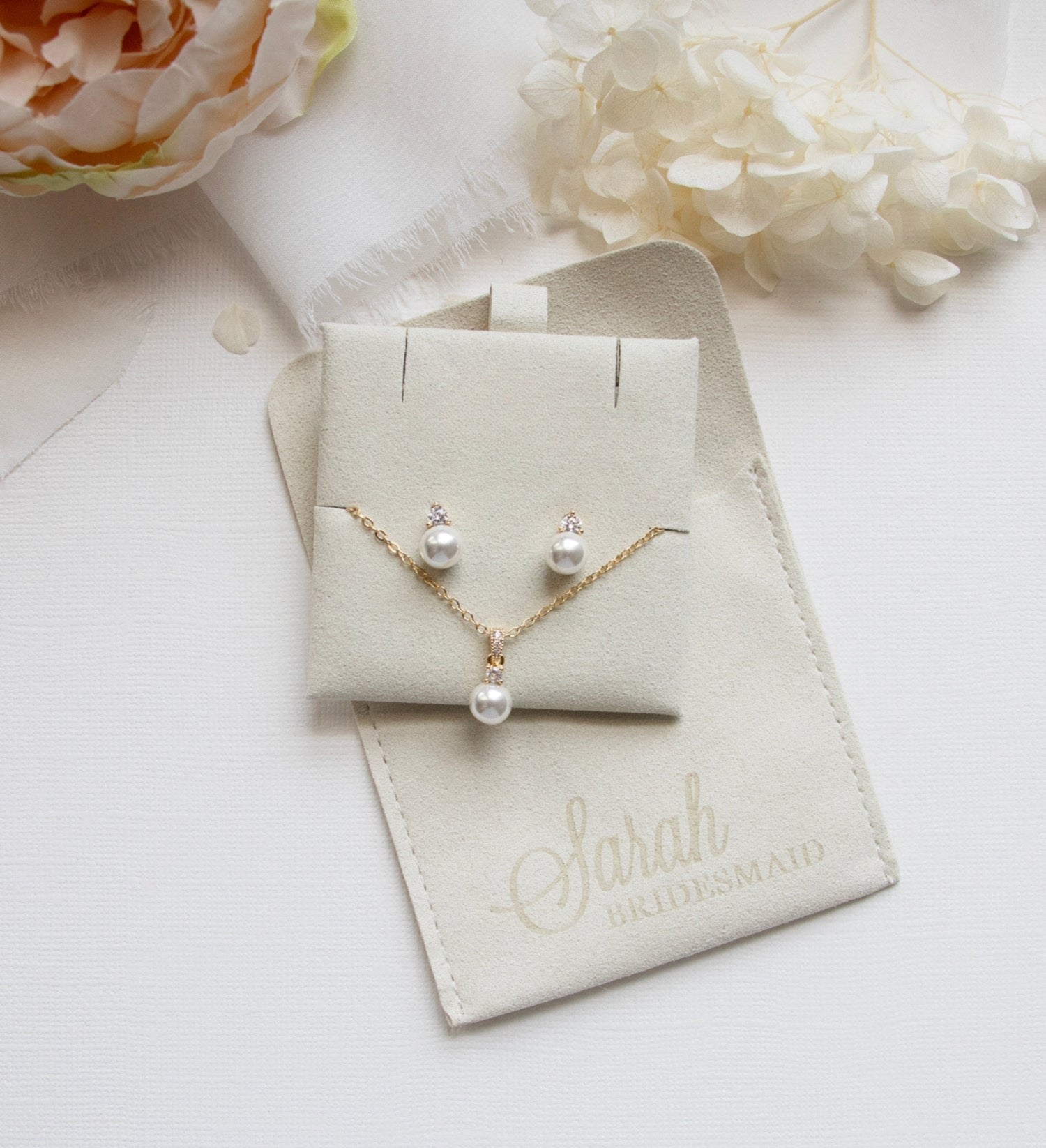 A necklace and earrings set are displayed on a white surface, accompanied by a small white flower and a beige pouch with the text "Sarah BRIDGETSmaid" printed on it.