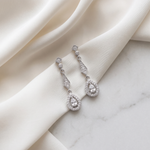 A pair of elegant diamond earrings with dangling designs rests on a white fabric surface.