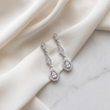 A pair of elegant diamond earrings with dangling designs rests on a white fabric surface.