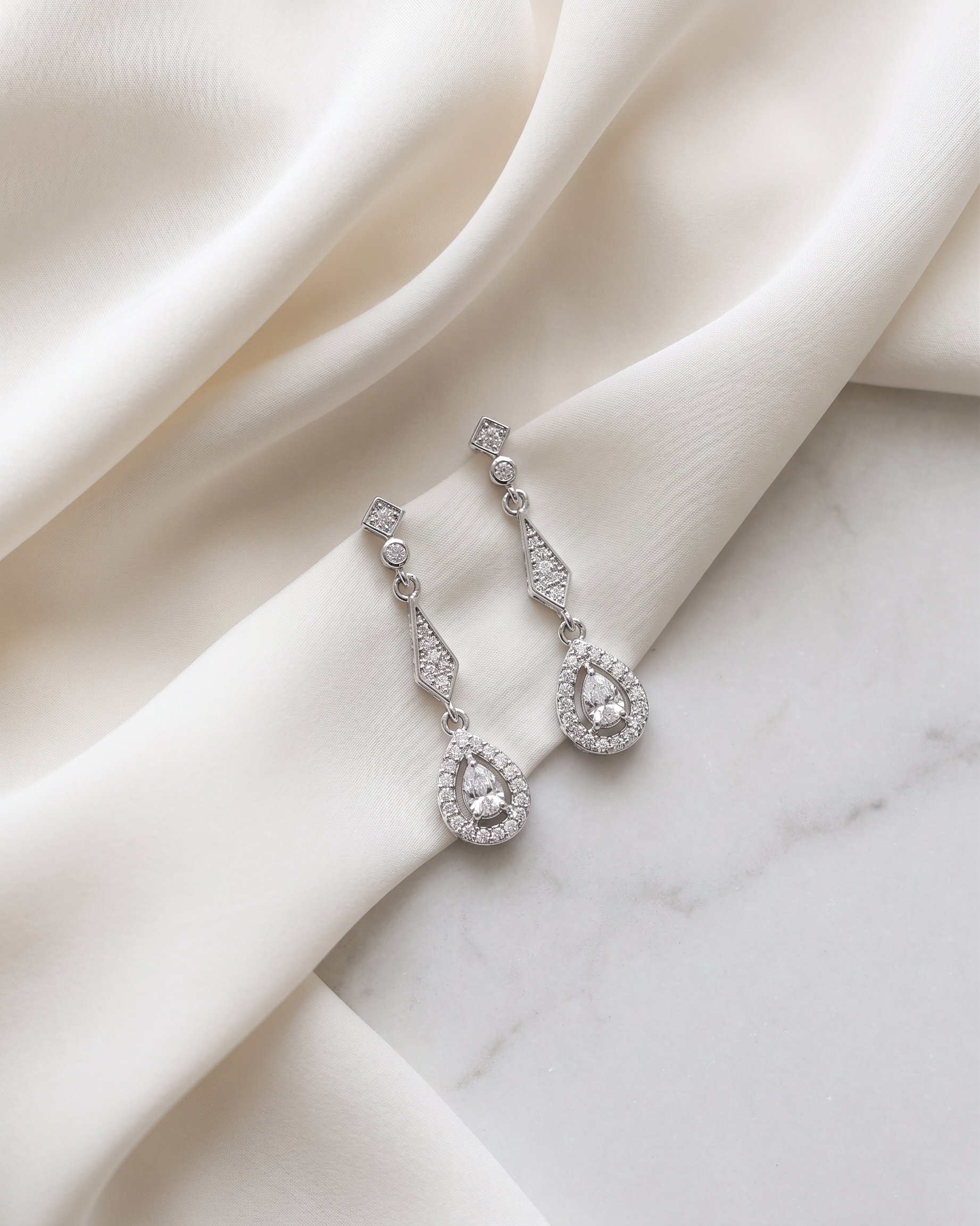 A pair of elegant diamond earrings with dangling designs rests on a white fabric surface.