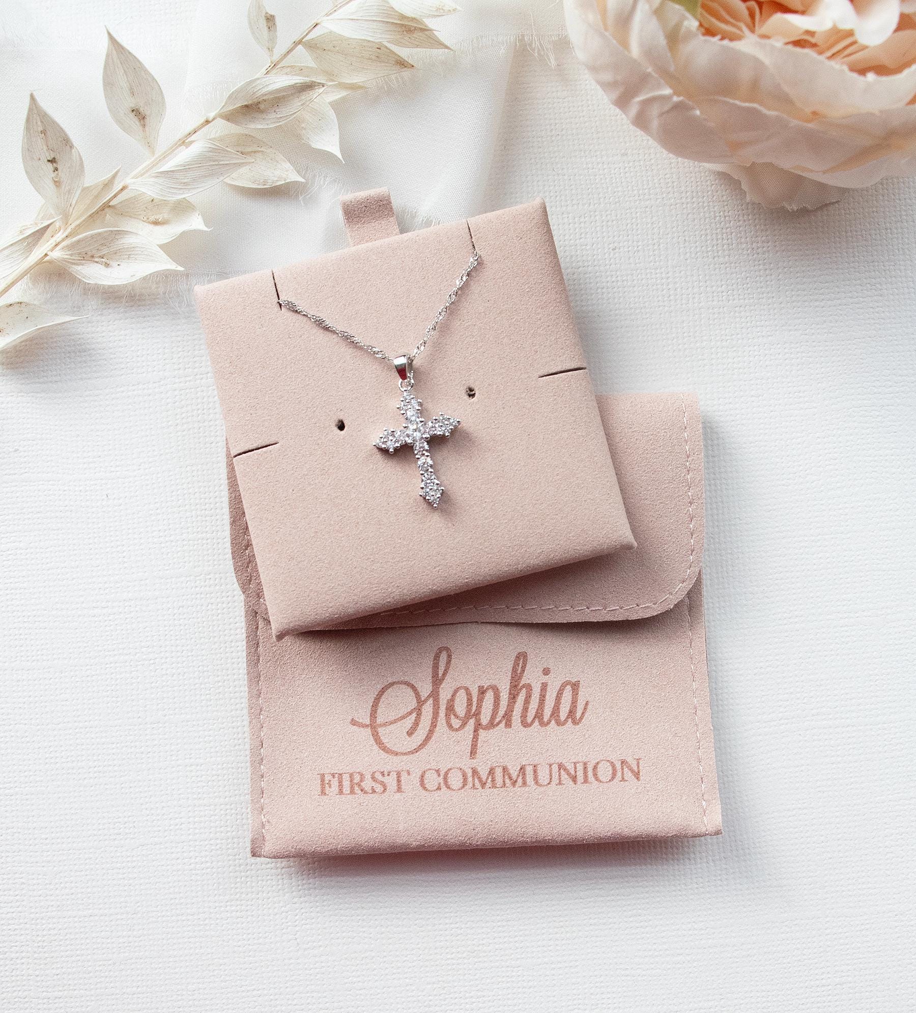 A necklace with a cross pendant is displayed in a beige box, accompanied by a small white flower and a personalized pouch with the name "Sophia" printed on it.