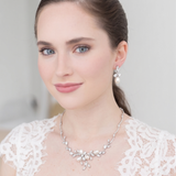 A woman wearing a white lace dress and a necklace with a floral design, smiling at the camera.