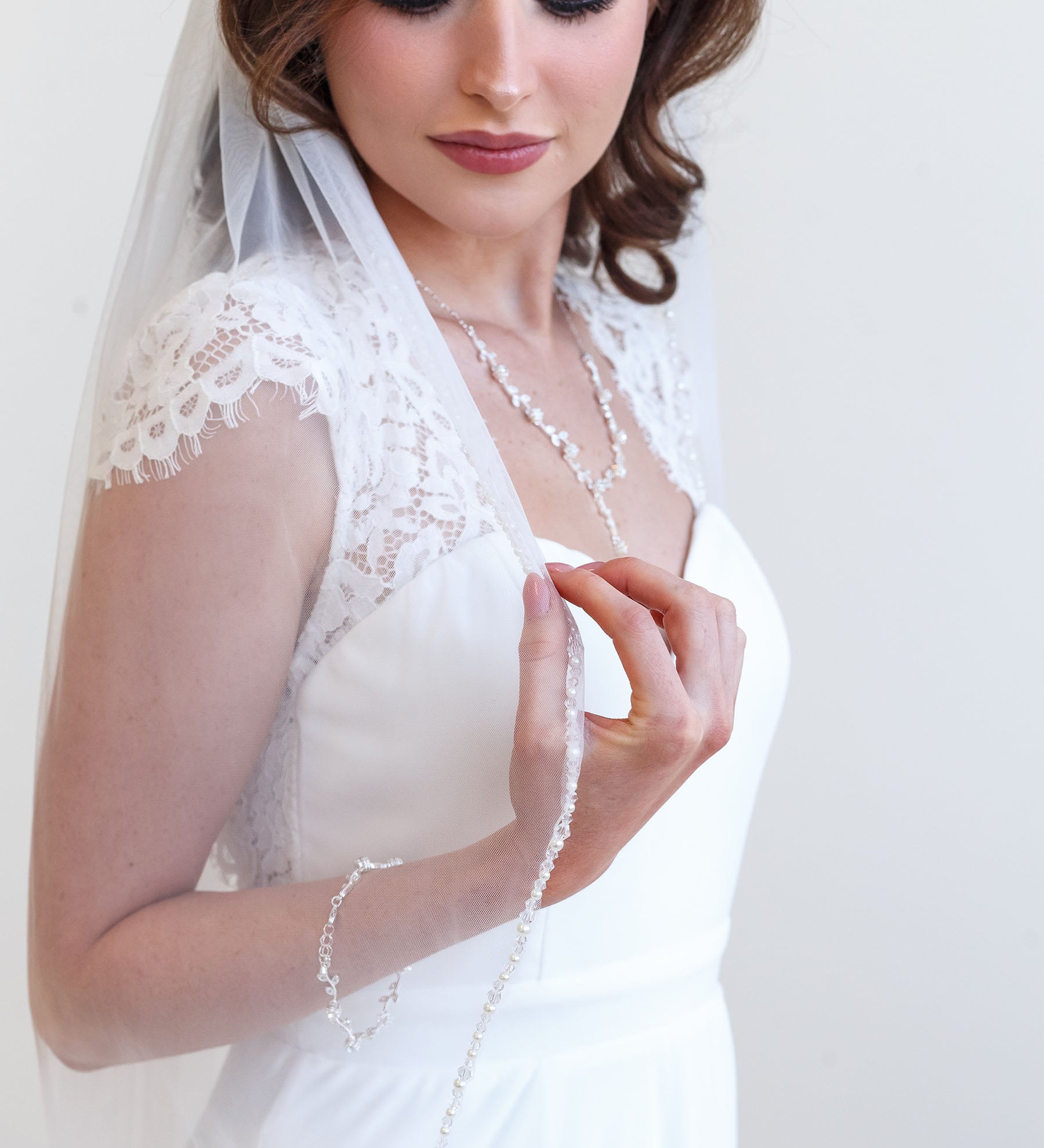 A woman in a white wedding dress with a veil and bracelet stands against a white background.