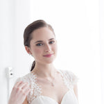 A young woman in a white lace dress stands in front of a white wall, smiling at the camera.
