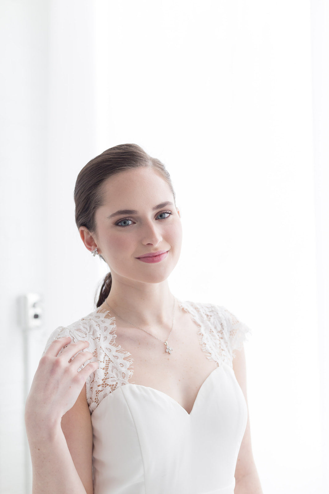 A young woman in a white lace dress stands in front of a white wall, smiling at the camera.
