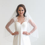 A woman wearing a white wedding dress with a veil, standing in front of a plain white background.