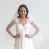 A woman wearing a white wedding dress with a veil, standing in front of a plain white background.