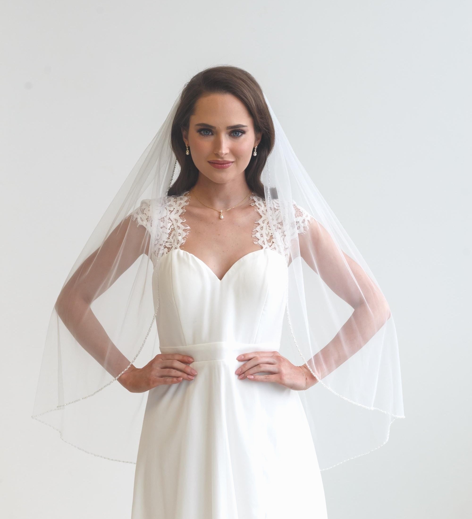 A woman wearing a white wedding dress with a veil, standing in front of a plain white background.
