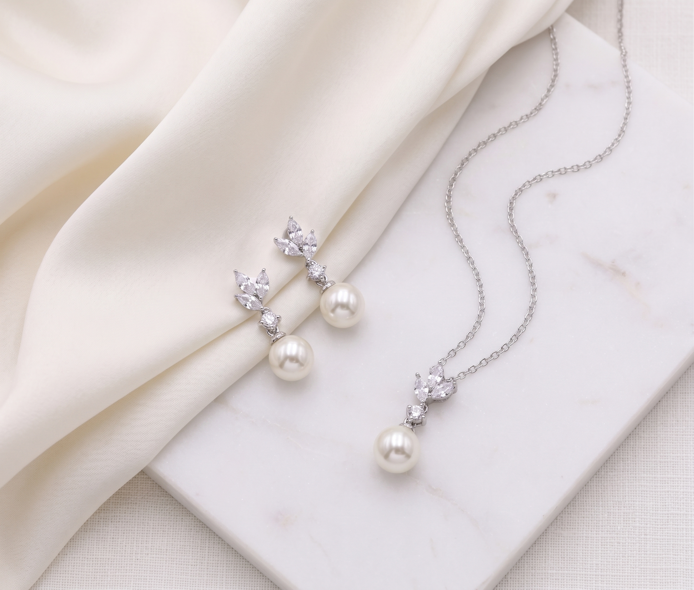 A pair of pearl earrings and a necklace with pearls are displayed on a white marble surface, with a white cloth draped over the background.