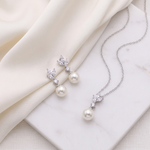 A pair of pearl earrings and a necklace with pearls are displayed on a white marble surface, with a white cloth draped over the background.