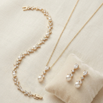 A delicate gold necklace, bracelet, and earrings are displayed on a beige fabric background.