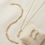 A delicate gold necklace, bracelet, and earrings are displayed on a beige fabric background.