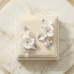 A pair of elegant white floral earrings with pearls and leaves, displayed on a white velvet box against a marble background.