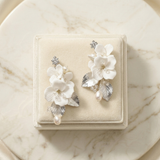 A pair of elegant white floral earrings with pearls and leaves, displayed on a white velvet box against a marble background.
