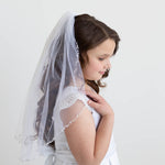 A young girl in a white dress with a veil stands against a plain white background, her gaze directed downwards.