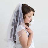 A young girl in a white dress with a veil stands against a plain white background, her gaze directed downwards.