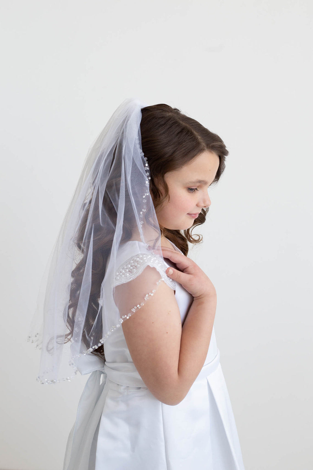 A young girl in a white dress with a veil stands against a plain white background, her gaze directed downwards.