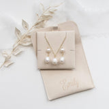 A beige jewelry box containing a necklace and earrings, with a personalized leather pouch labeled "Emilly" placed next to it.