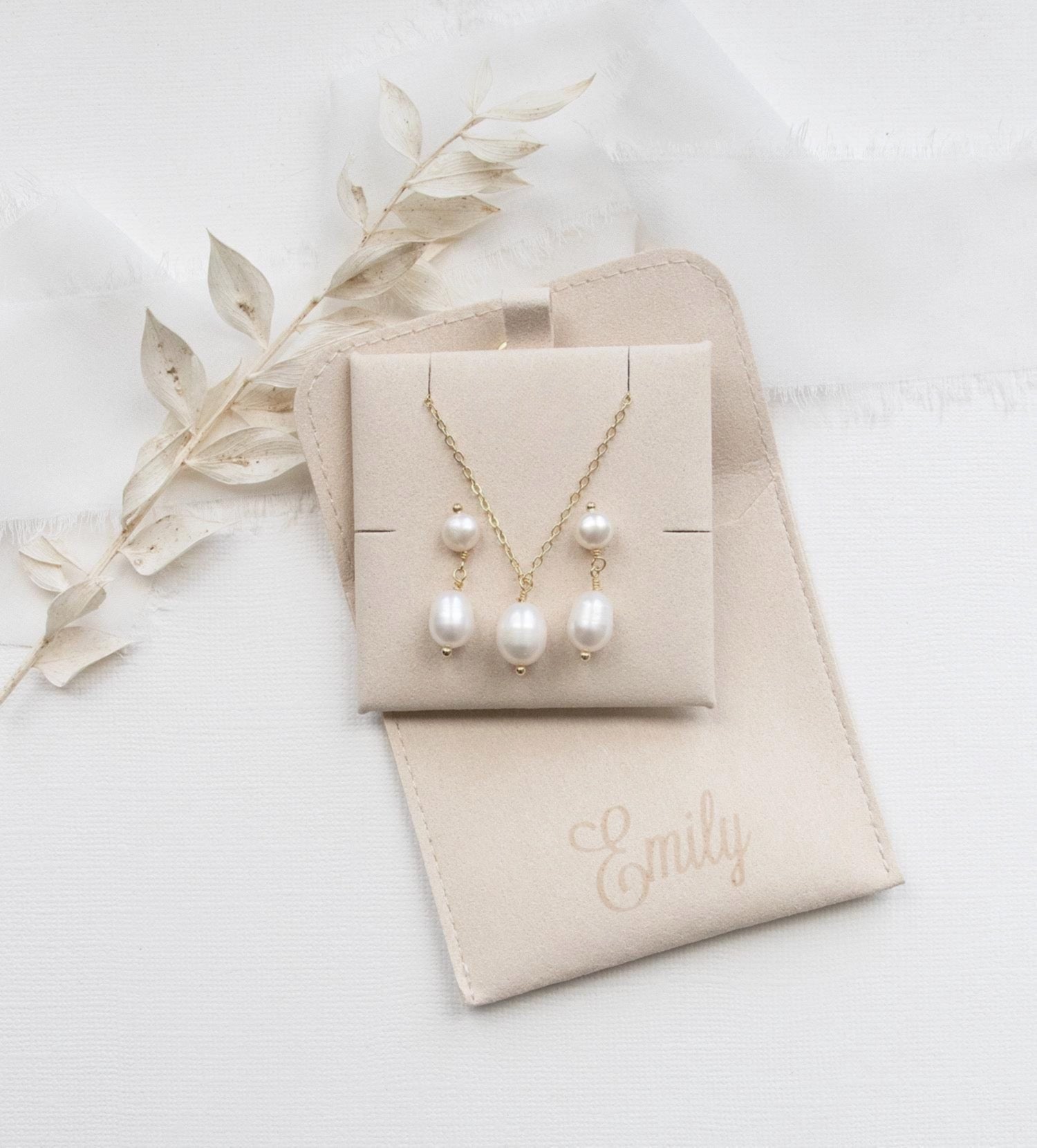 A beige jewelry box containing a necklace and earrings, with a personalized leather pouch labeled "Emilly" placed next to it.