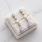 A pair of elegant earrings with pearls and diamonds displayed in a white jewelry box on a marble surface.