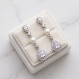 A pair of elegant earrings with pearls and diamonds displayed in a white jewelry box on a marble surface.