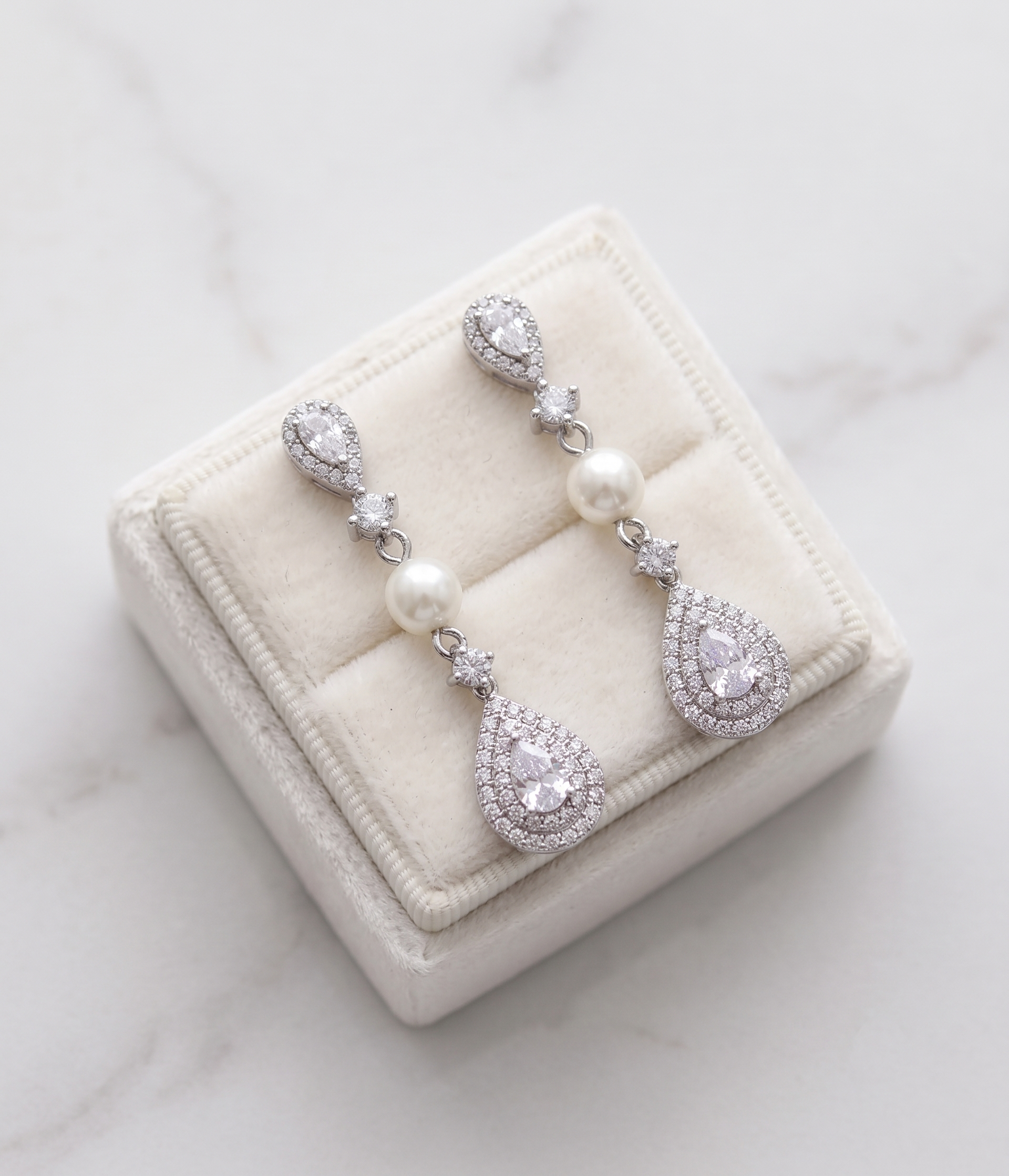 A pair of elegant earrings with pearls and diamonds displayed in a white jewelry box on a marble surface.