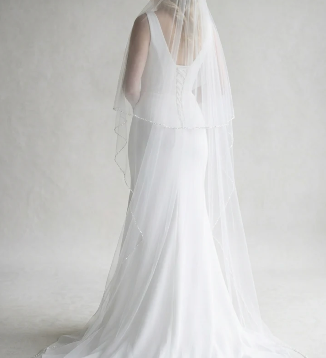 A woman in a long white wedding dress with a veil stands facing away from the camera, her back to the viewer.
