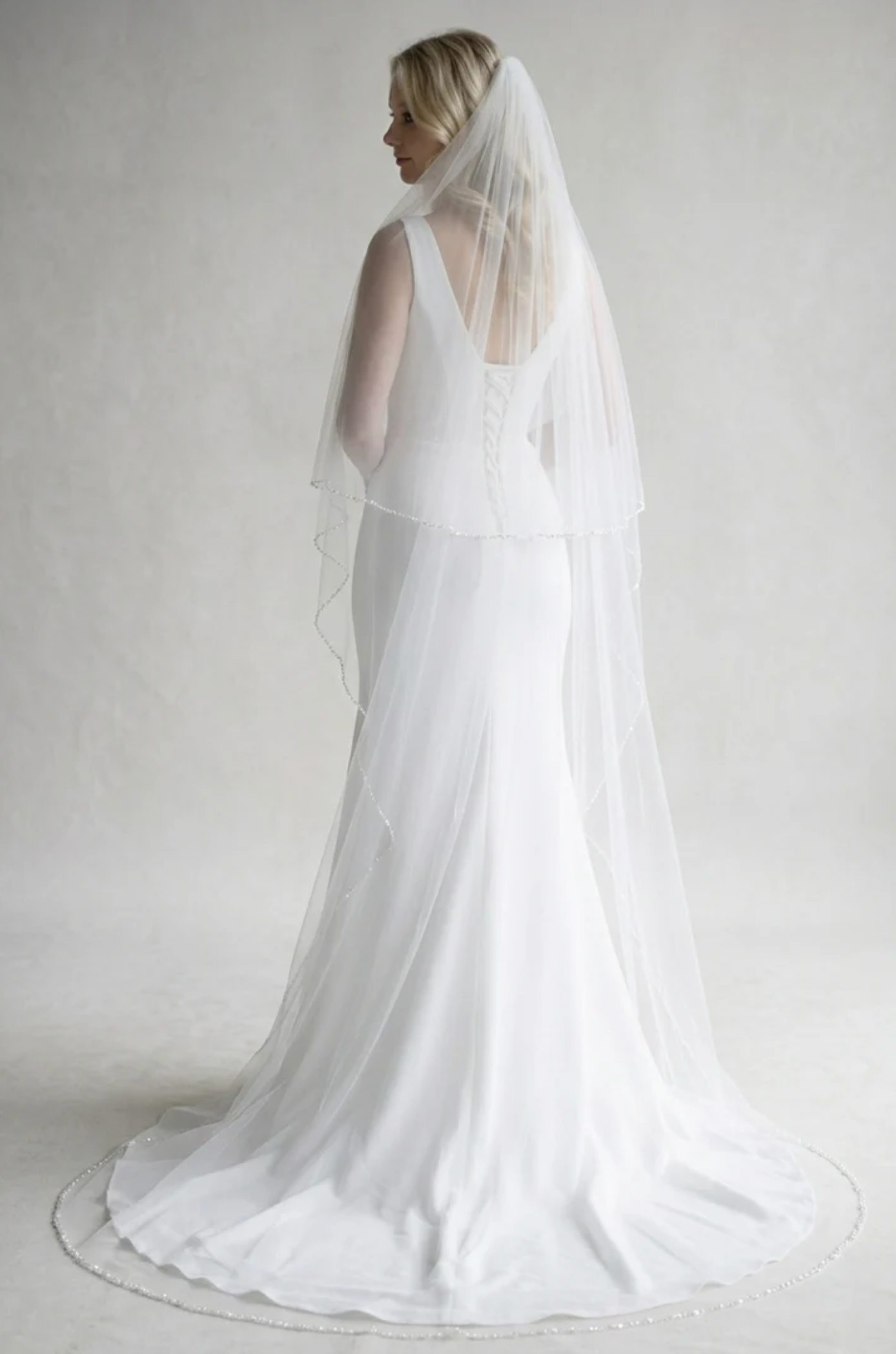 A woman in a long white wedding dress with a veil stands facing away from the camera, her back to the viewer.