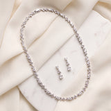 A necklace and earrings are displayed on a white marble surface, with the necklace featuring a delicate chain of small, sparkling diamonds.
