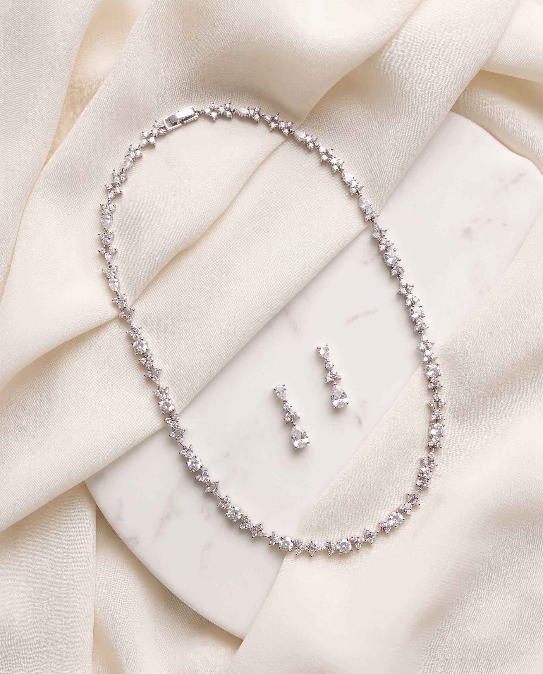 A necklace and earrings are displayed on a white marble surface, with the necklace featuring a delicate chain of small, sparkling diamonds.