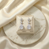 A pair of elegant diamond earrings is displayed on a white marble surface, placed in a small white box.