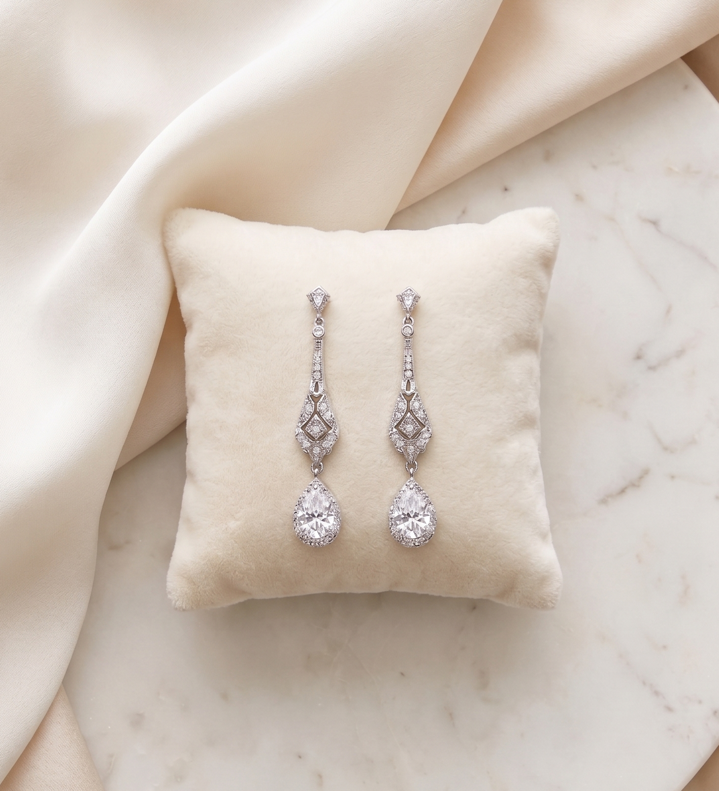 A pair of elegant diamond earrings with dangling designs rests on a white pillow against a beige background.