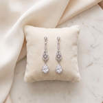A pair of elegant diamond earrings with dangling designs rests on a white pillow against a beige background.