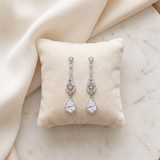 A pair of elegant diamond earrings with dangling designs rests on a white pillow against a beige background.