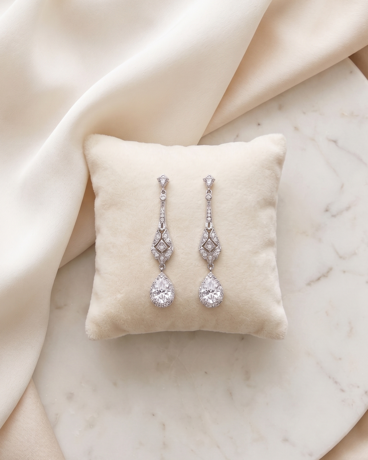 A pair of elegant diamond earrings with dangling designs rests on a white pillow against a beige background.