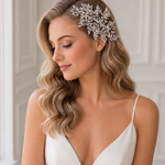 A woman with long, wavy blonde hair wearing a white dress and a decorative headpiece stands in front of a white wall.