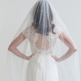 A woman wearing a white wedding dress with a veil, standing against a white background.