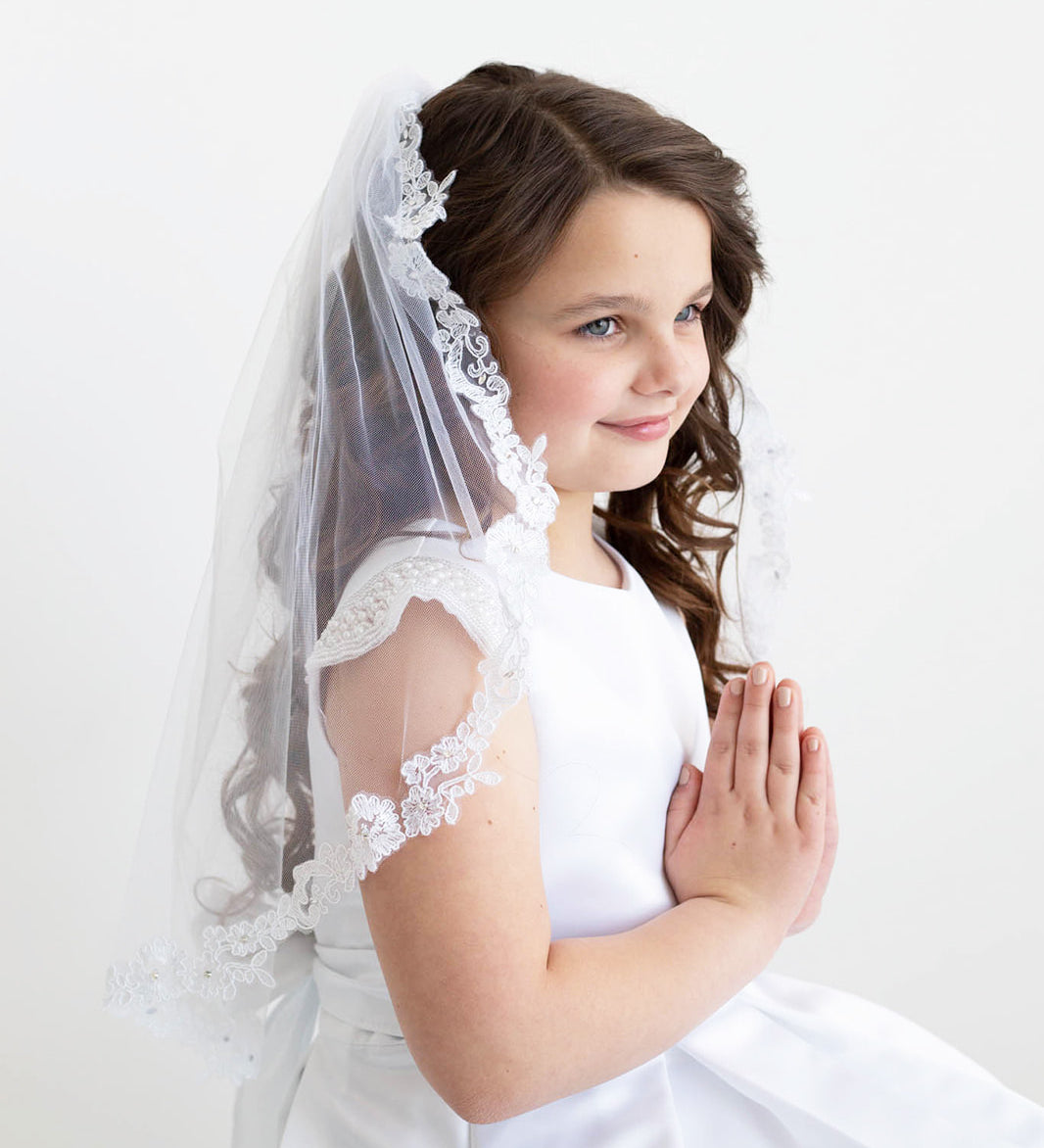 A young girl in a white dress and veil stands against a plain white background, her hands clasped together in a prayer position.