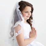 A young girl in a white dress and veil stands against a plain white background, her hands clasped together in a prayer position.