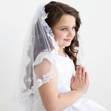 A young girl in a white dress and veil stands against a plain white background, her hands clasped together in a prayer position.