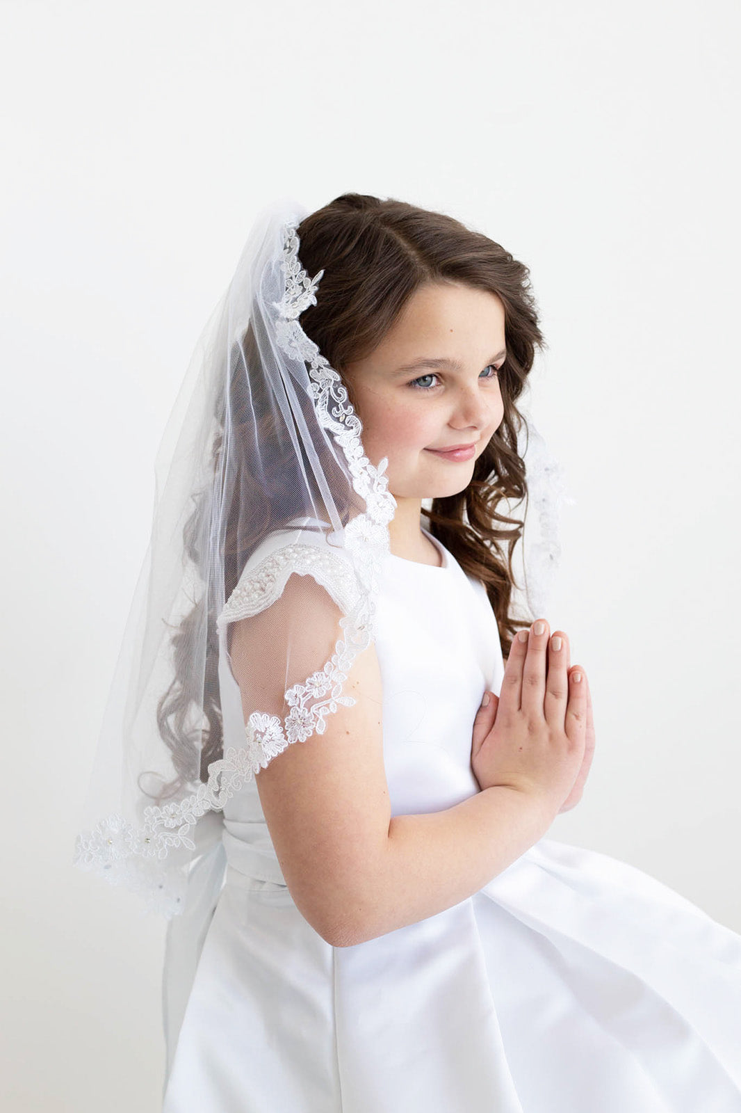 A young girl in a white dress and veil stands against a plain white background, her hands clasped together in a prayer position.