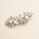 A decorative hair clip adorned with numerous sparkling crystal elements, including pearls and diamonds, set against a light beige background.