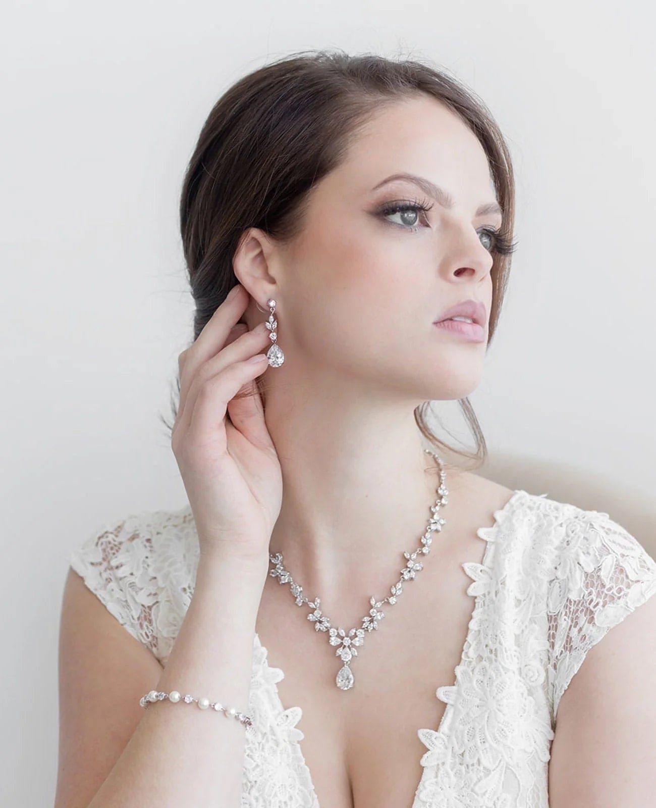 A woman in a white lace dress and diamond jewelry poses against a plain white background.