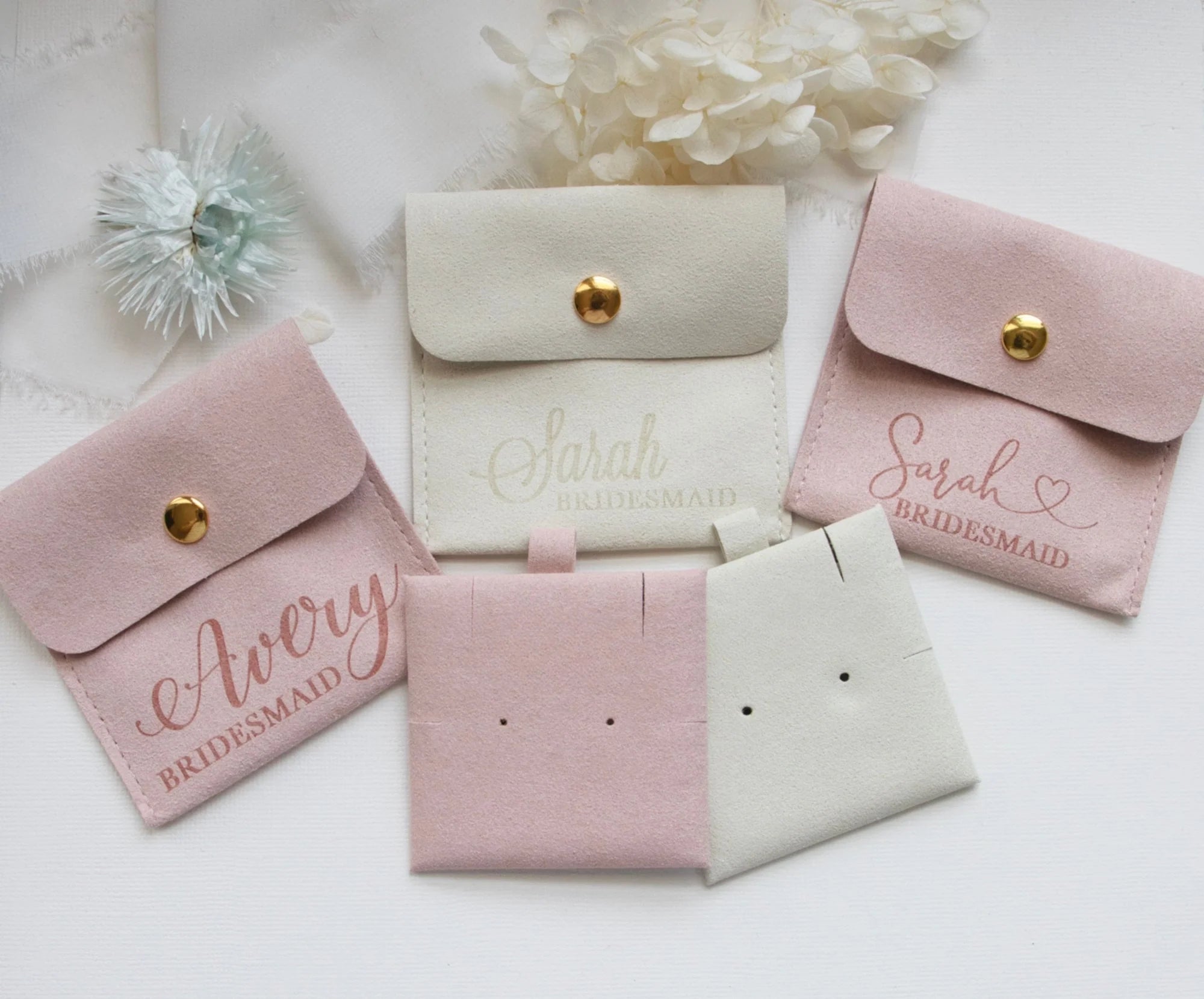 a collection of pink and white bridesmaid bags with personalized names on them, along with some white flowers and a white fabric background.