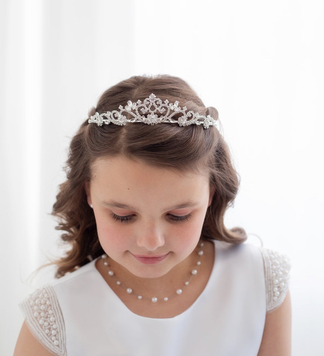 A young girl with a tiara on her head, wearing a white dress and pearl necklace, looking down at the ground.