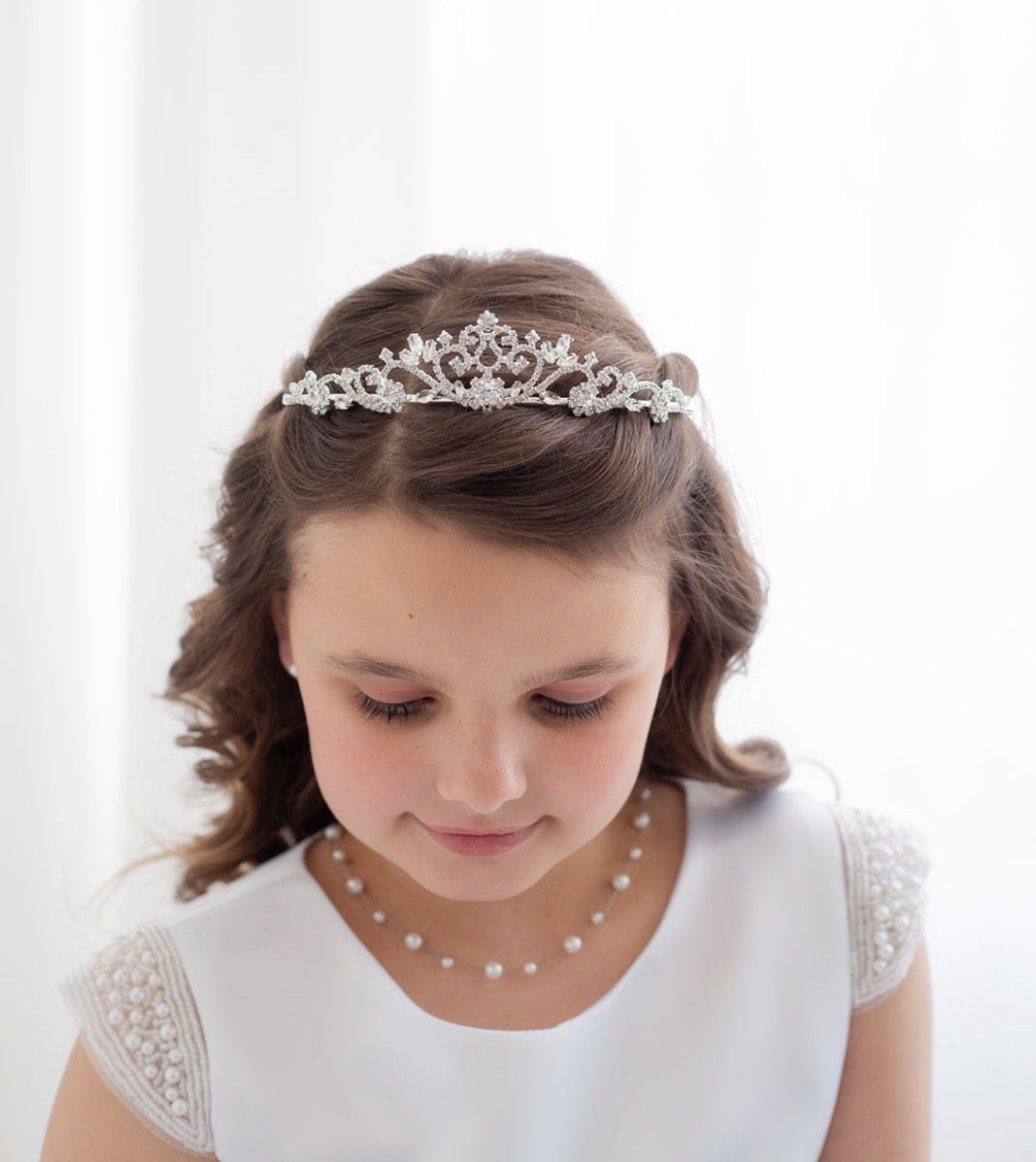 A young girl with a tiara on her head, wearing a white dress and pearl necklace, looking down at the ground.