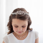 A young girl with a tiara on her head, wearing a white dress and pearl necklace, looking down at the ground.