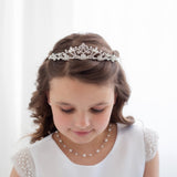 A young girl with a tiara on her head, wearing a white dress and pearl necklace, looking down at the ground.