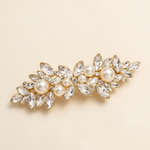 A decorative hair clip adorned with numerous sparkling crystal elements, including pearls and diamonds, set against a light beige background.